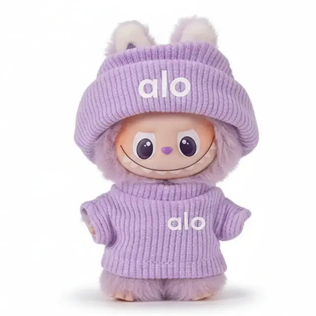 Doll habillée en violet avec le mot "alo" sur le chandail et le bonnet, ajoutant une touche ludique à la collection.